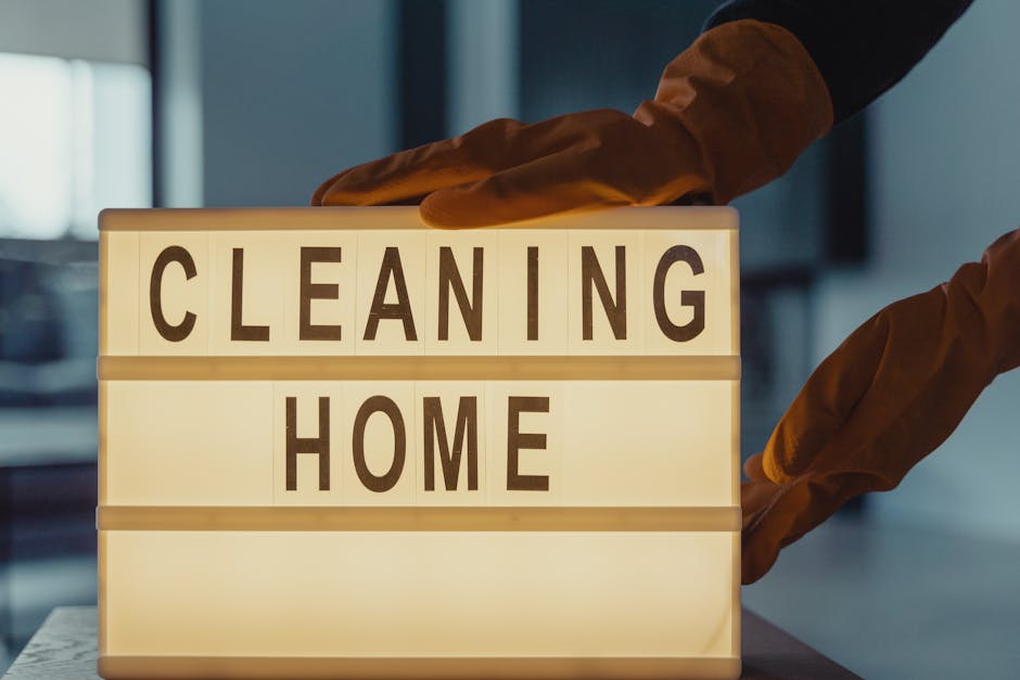 A person wearing brown cleaning gloves is positioning a lightbox sign that reads 'CLEANING HOME' in bold black letters on a smooth surface. The background features a dimly lit, modern interior with a