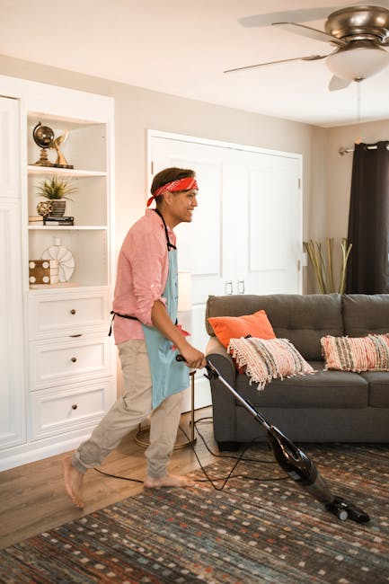 A woman performing surface cleaning on a patterned beige and cream area rug using a yellow vacuum cleaner with a hose attachment. She is standing on the rug in a living room, wearing casual clothing w